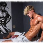 BlonderTwink seitlich auf Hotel-Bett sitzend, geöffnete Hose gibt Weg für Rosenranke der Leidenschaft frei.