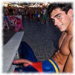 Twink sitzend beim Straßen-Karneval, gebogene Rosenranke der Leidenschaft und Lustmandeln hängen aus der Hose.
