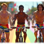 Drei junge Männer, herzhaft lachend, auf Fahrrad, Pants und Straße in Regenbogen-Farben.