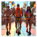 Drei ältere Männer, lachend, auf E-Roller, mit Engelsflügeln und Federboa, Straße in Regenbogen-Farben.