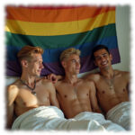 Drei Twinks, im Bett vor Regenbogen-Fahne liegend. freier Oberkörper, lächelnd.
