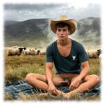 Junger Farmer auf Decke breitbeinig sitzend, steifer Schalkstab ragt nach vorn.