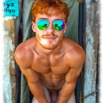 Rothaariger Twink mit Sonnenbrille und 3-Tage-Bart, Lustpendel leicht zwischen den Oberschenkeln eingeklemmt.